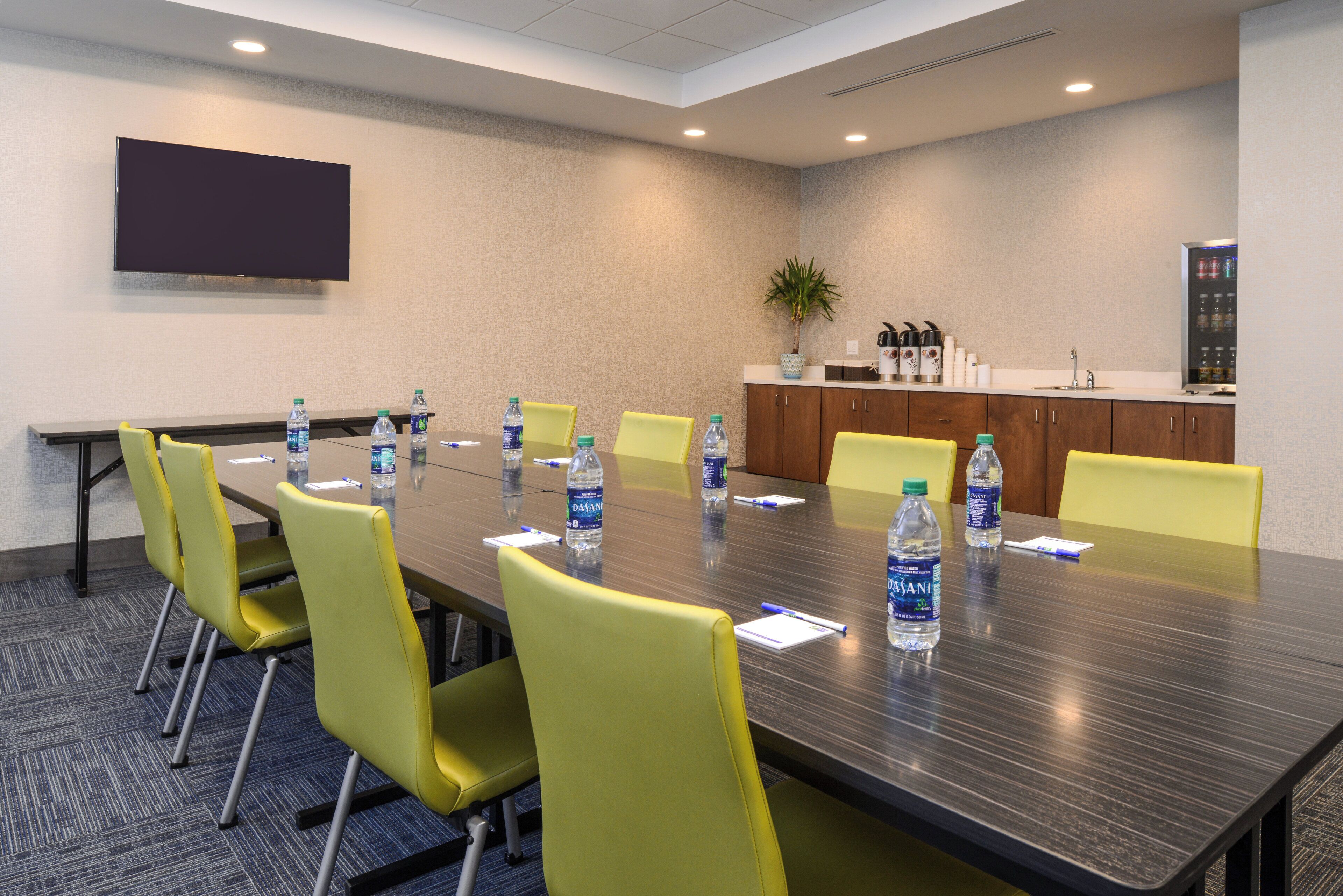A conference room with a long table, yellow chairs, water bottles, a TV, and a kitchenette.