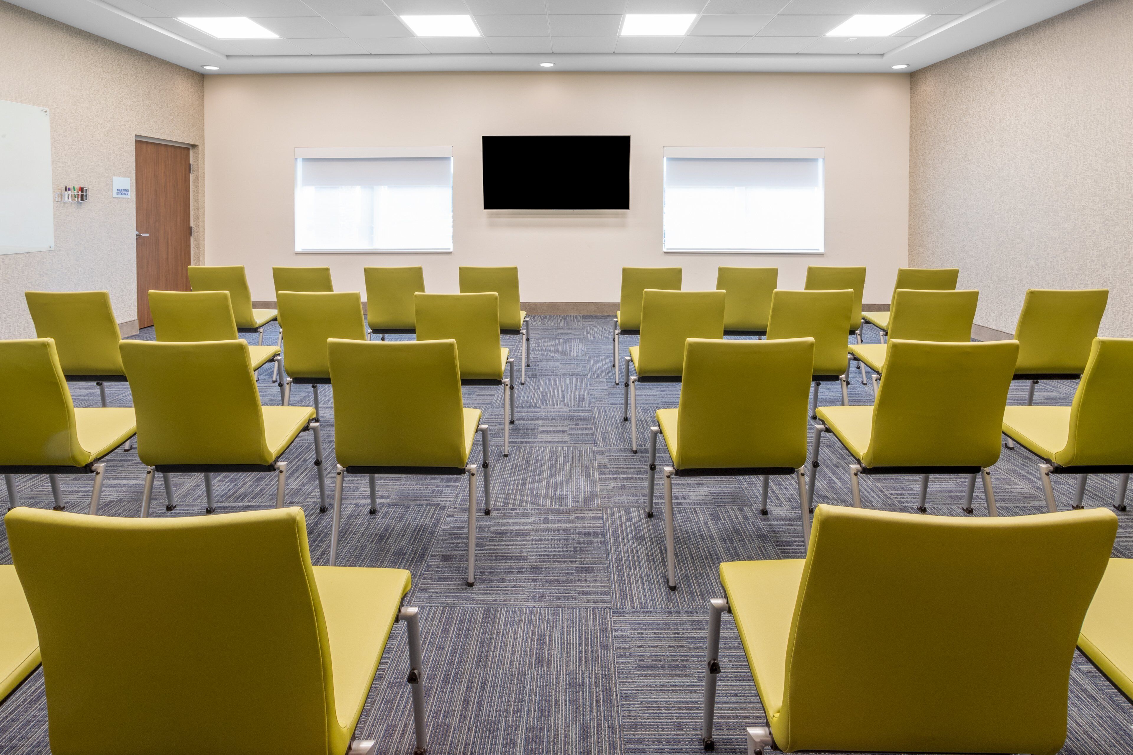 A conference room with rows of yellow chairs, a large screen, and two windows.