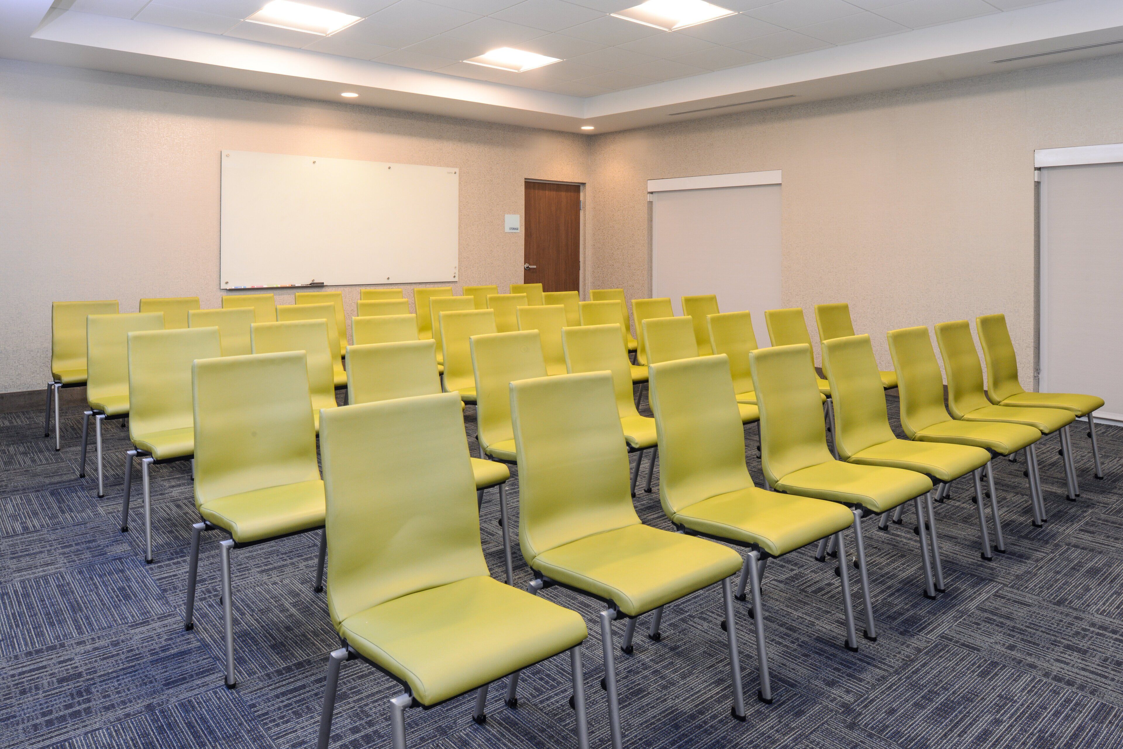 Une salle de conférence avec des rangées de chaises jaunes, un tableau blanc et une porte.