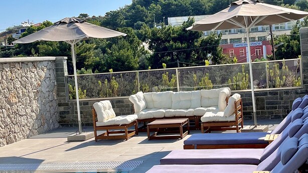 A poolside area with lounge chairs and a sofa, a stone wall, and a view of a hill with buildings in the background. - Villa Divina (Rhodes)