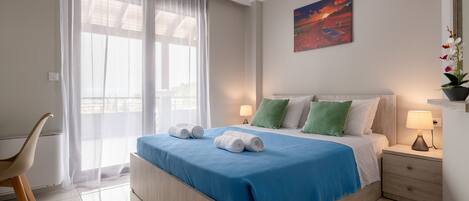 Een net opgemaakt bed met een blauwe deken en twee opgerolde handdoeken, een nachtkastje met een lamp, een stoel en een raam met dunne gordijnen.