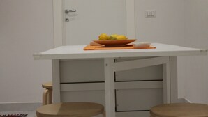 Una pequeña mesa blanca con un frutero y una taza, dos taburetes de madera, y una puerta blanca al fondo.