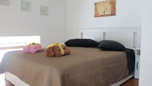 Una cama bien tendida con una colcha marrón y dos almohadas negras, una mesita de noche blanca y una pared con tres mapas enmarcados.