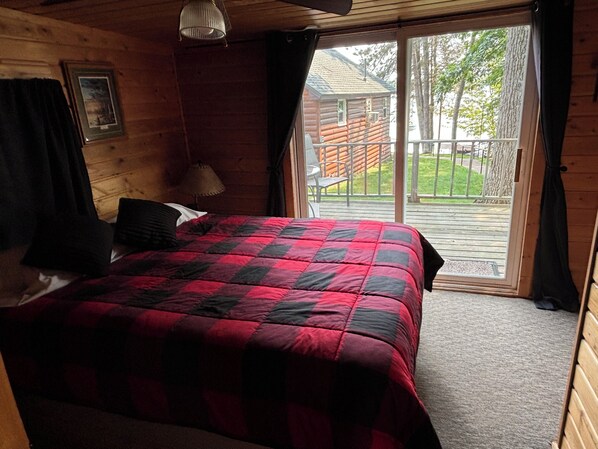 A wooden cabin room with a bed, a lamp, a window, and a view of a wooden deck and trees.