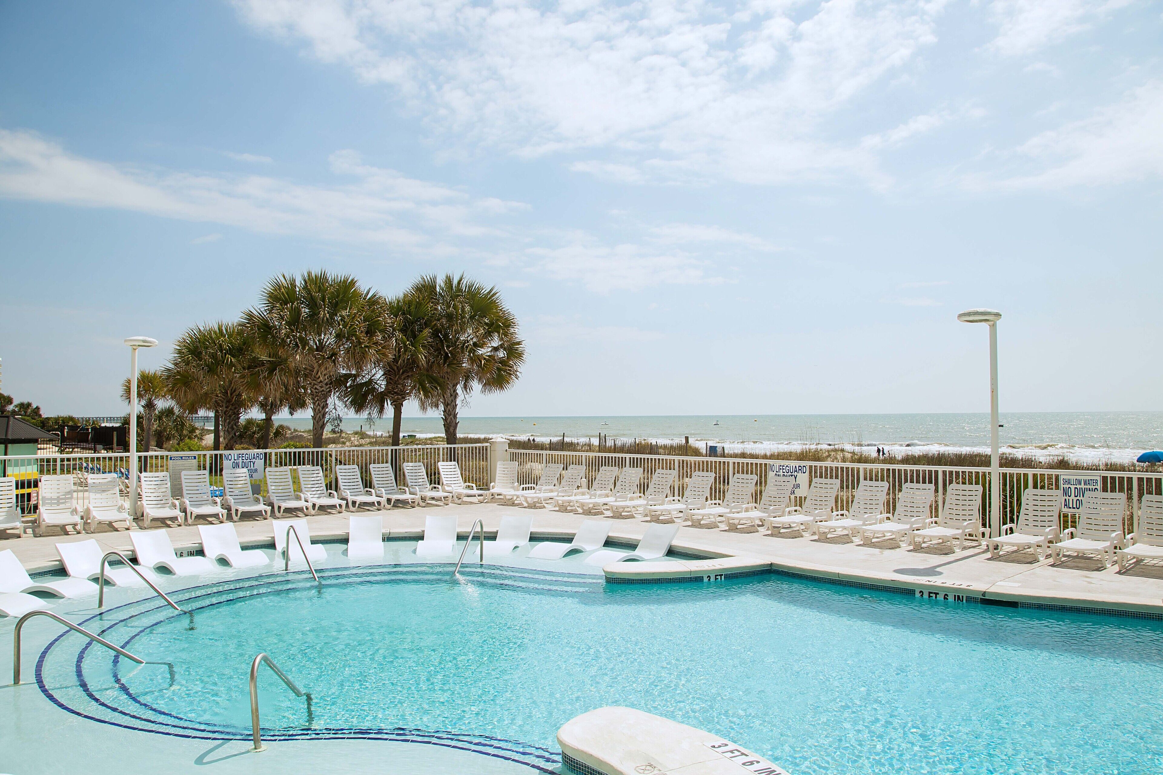 A swimming pool with lounge chairs, palm trees, and a view of the ocean.