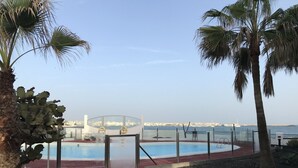 Área da piscina com estrutura de vidro, palmeiras e vista para o mar e a cidade.