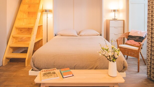 Un dormitorio con techo de madera, una cama, una mesita de noche, una silla y un escritorio con libros y un jarrón de flores.