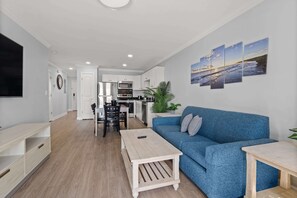 A modern living room with a blue sofa, a wooden coffee table, and a TV on the left. A kitchen is visible in the background with white cabinets and a microwave. - Premium Ocean View Two Bedroom Condo - FREE Pickleball 5 minutes away! (Myrtle Beach)