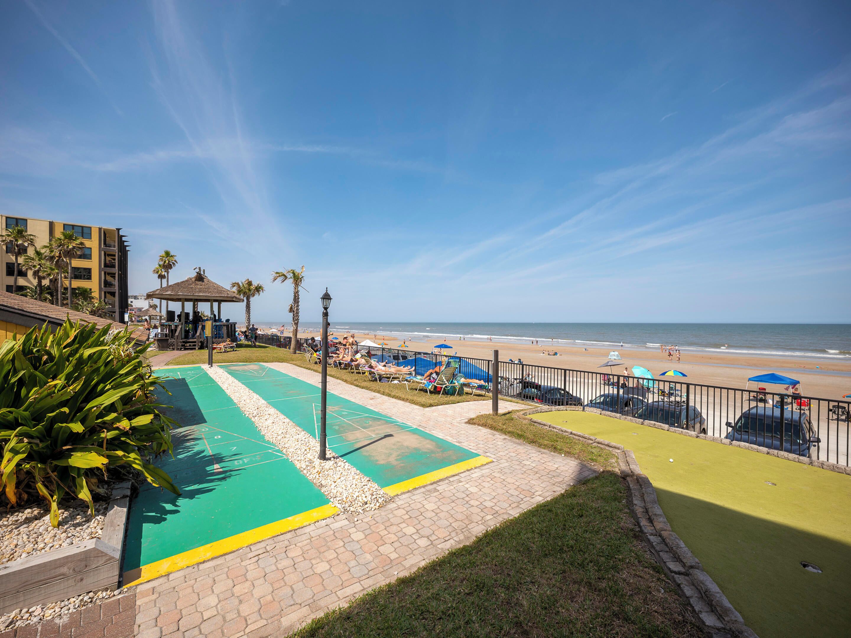 A beachfront view with a green shuffleboard court, palm trees, and beach umbrellas.