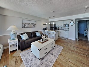 A modern living room with a sofa, coffee table, and a dining area with chairs and a kitchen in the background.