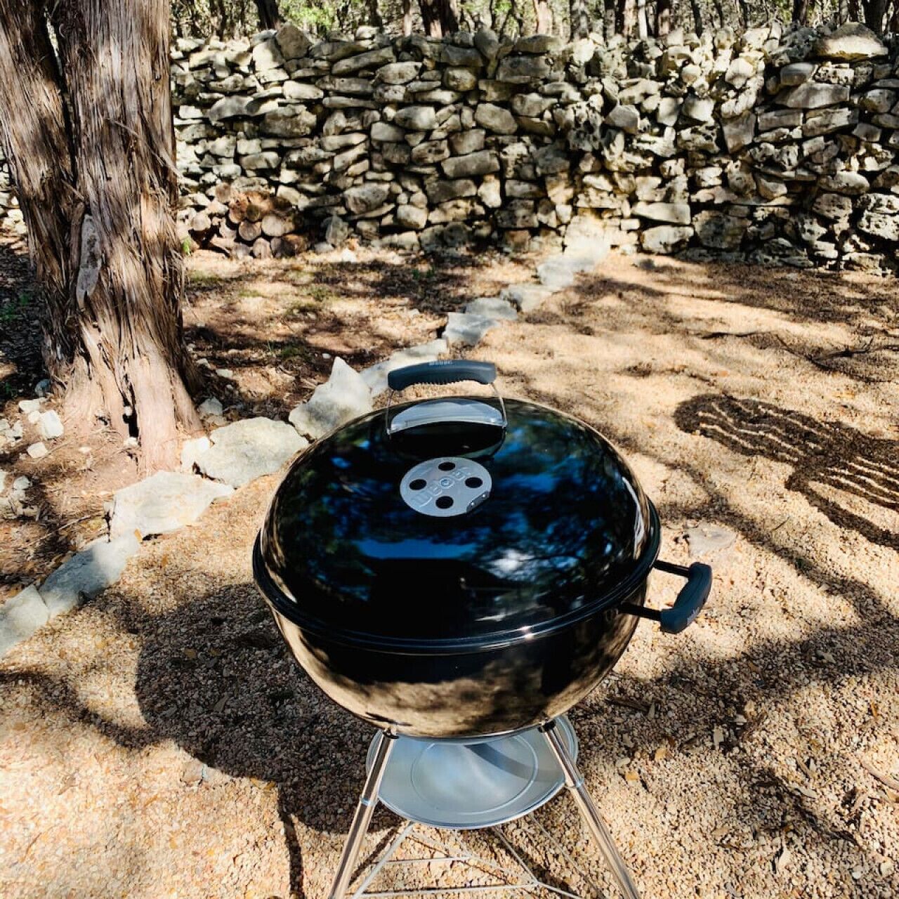 A portable barbecue grill with a lid, set on a metal stand, outdoors with a stone wall and tree in the background.