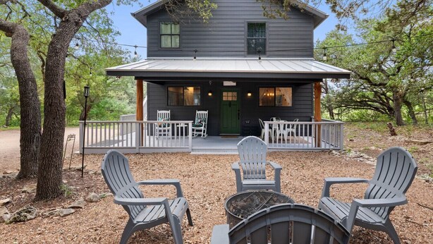 A two-story cabin with a porch, outdoor seating, and a large tree in the foreground. - Cottage: Fire Pit | Covered Porch | Sleeps 4 | Wimberley TX (Wimberley)