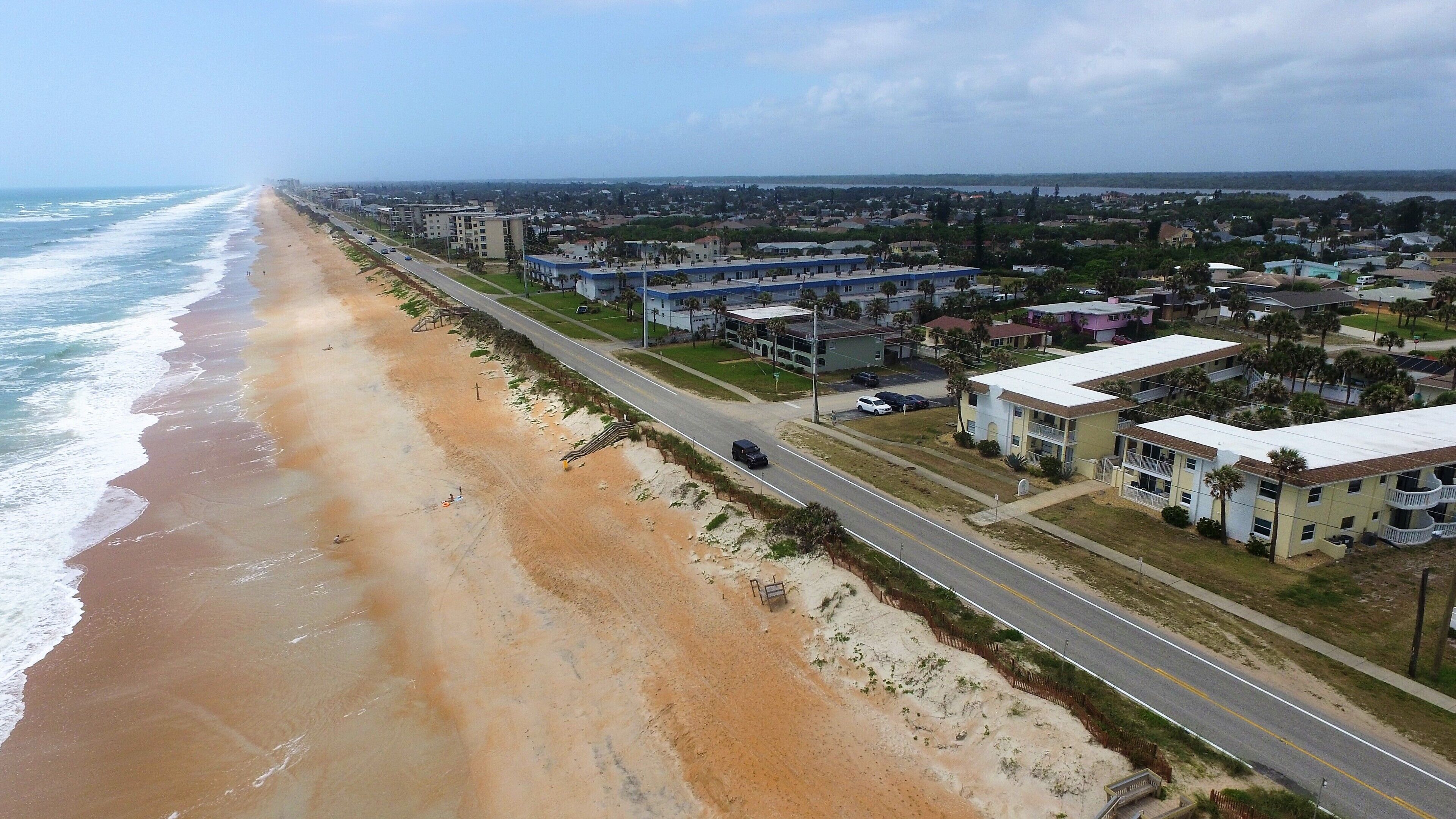 An aerial view of a coastal town with a beach, a road, and residential buildings.