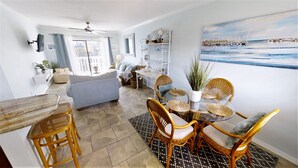 A spacious living area with a sofa, dining table with wicker chairs, a ceiling fan, and a large window. - OCEAN VIEW CONDO w/ Pool, Cable, Wifi, BEACH ACCESS. Ormond-by-the-Sea  (Ormond Beach)