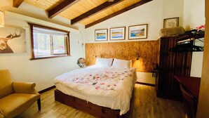 Une chambre avec un plafond en bois, un lit recouvert d’une literie à motifs floraux, un banc en bois, une chaise et une fenêtre donnant sur des arbres.