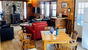L’intérieur chaleureux d’un chalet avec une cheminée en pierre, des murs en bois, un coin repas équipé d’un ordinateur portable et un canapé rouge.
