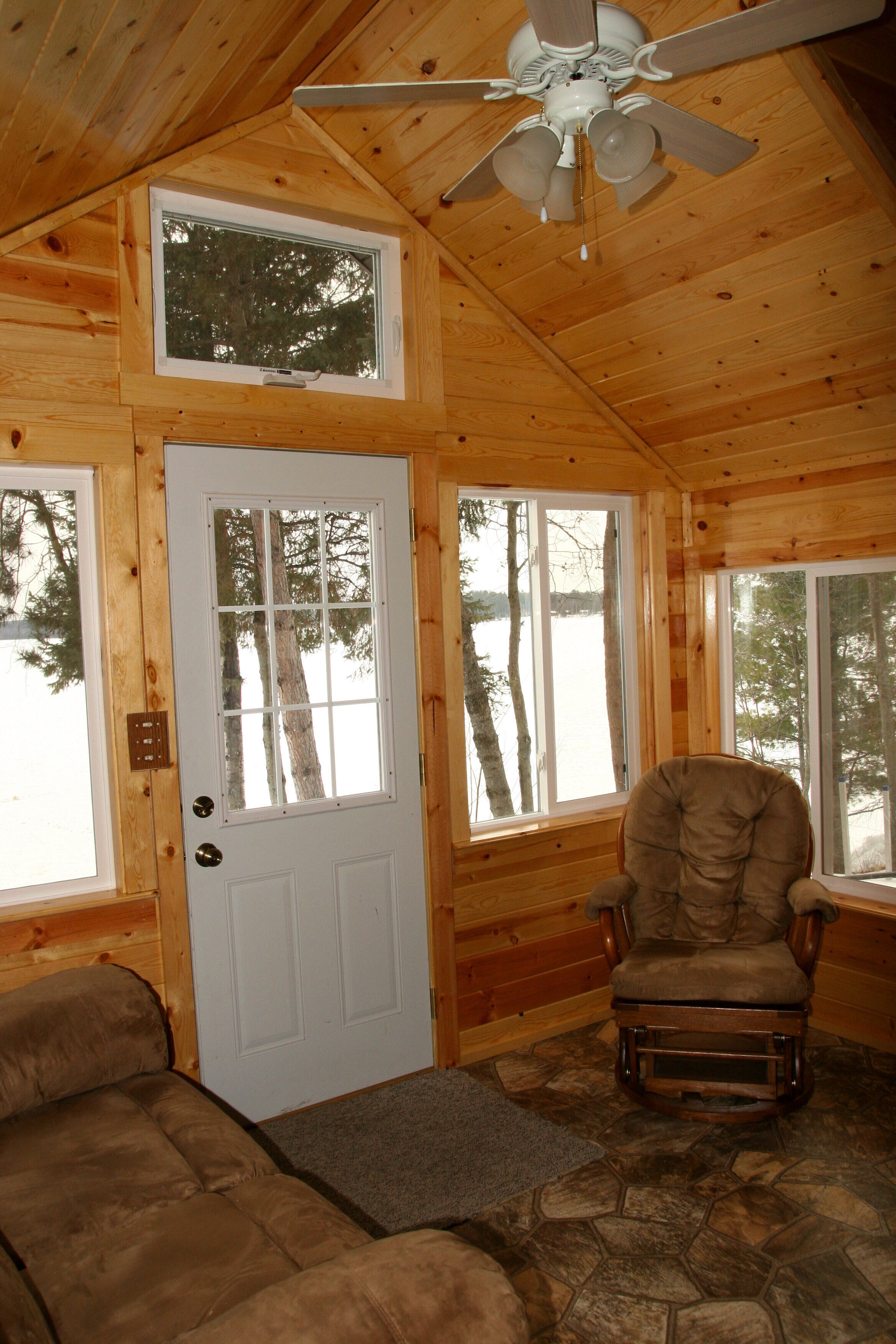 A log cabin interior with a wooden ceiling, a ceiling fan, a door with a window, a rocking chair, and a stone-patterned floor.