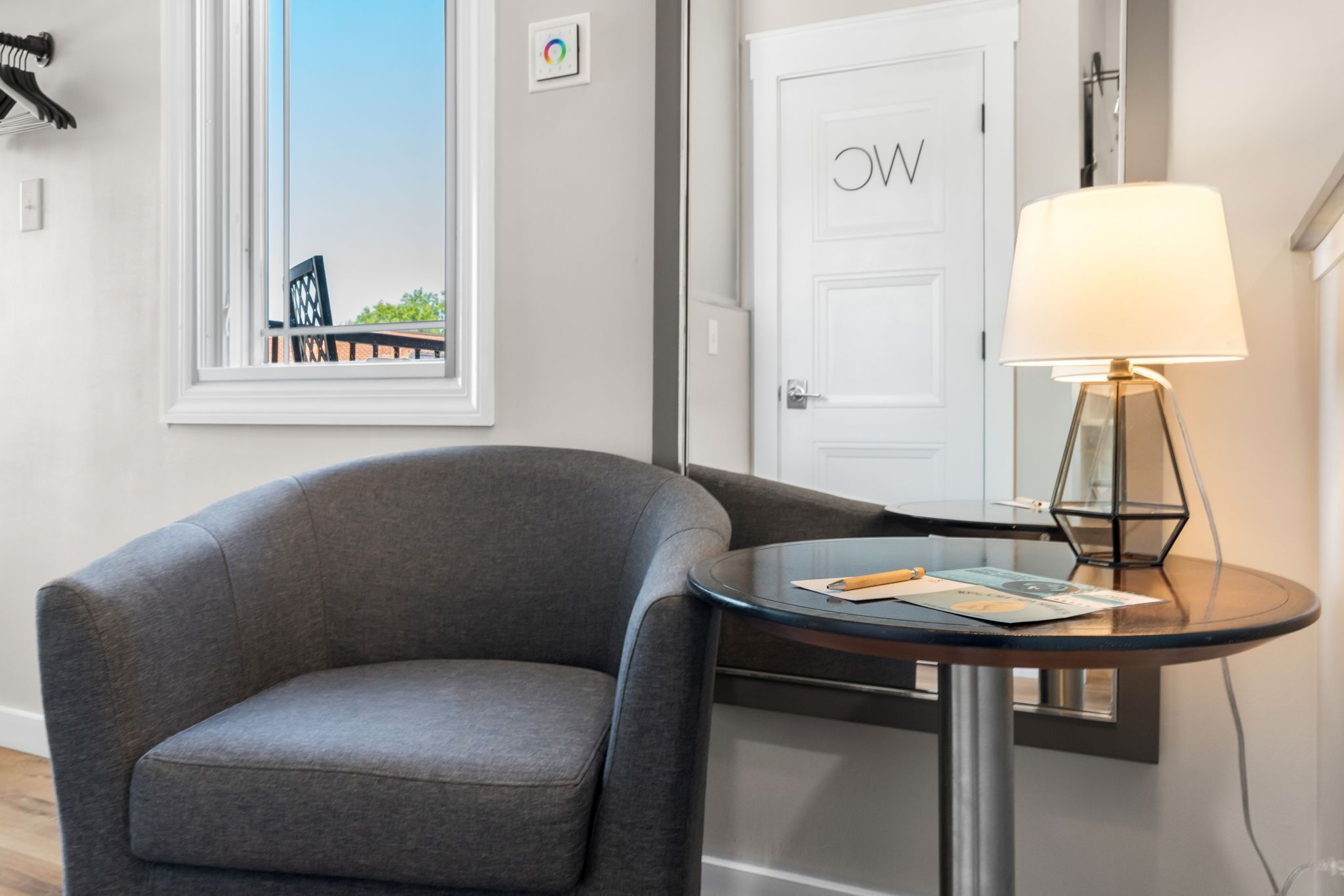 A modern room with a grey armchair, a round table with a lamp, and a door with a 'W' sign.