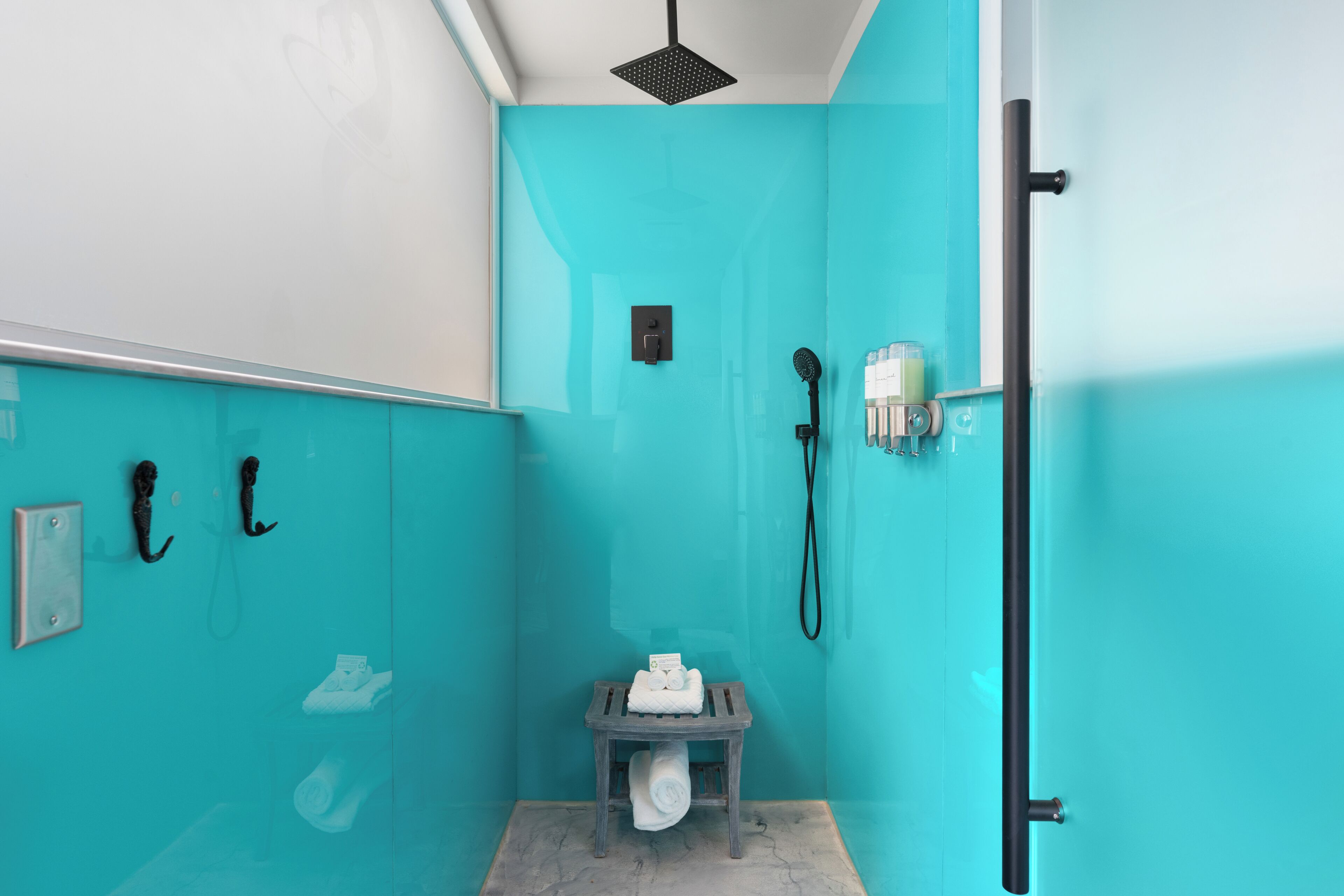 A bathroom with a turquoise wall, a shower with a handheld showerhead, a towel rack, and a small wooden stool.