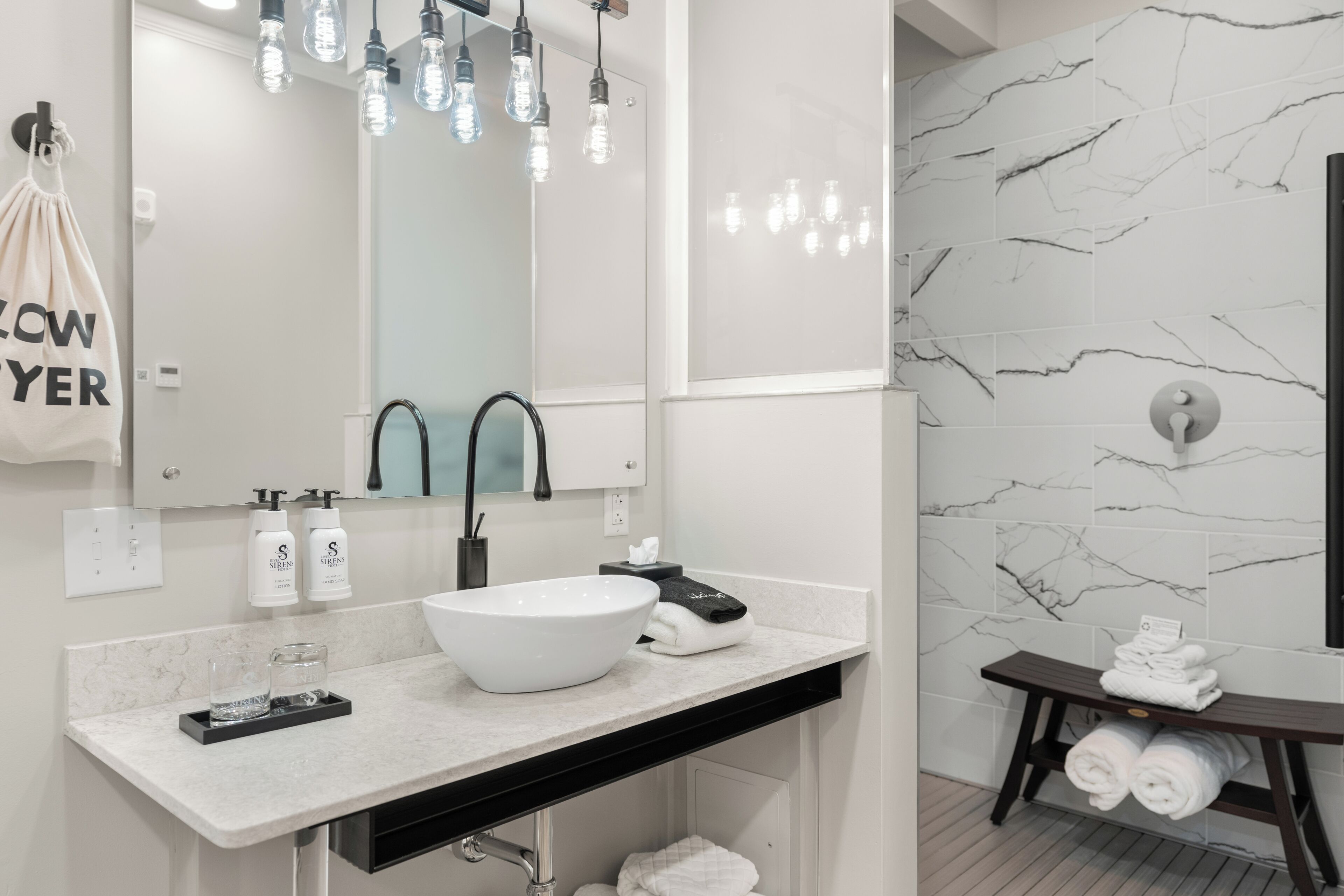 Une salle de bain moderne avec un lavabo blanc, un robinet noir et un pare-baignoire en marbre. On y voit deux flacons de savon liquide et une serviette accrochée au mur.