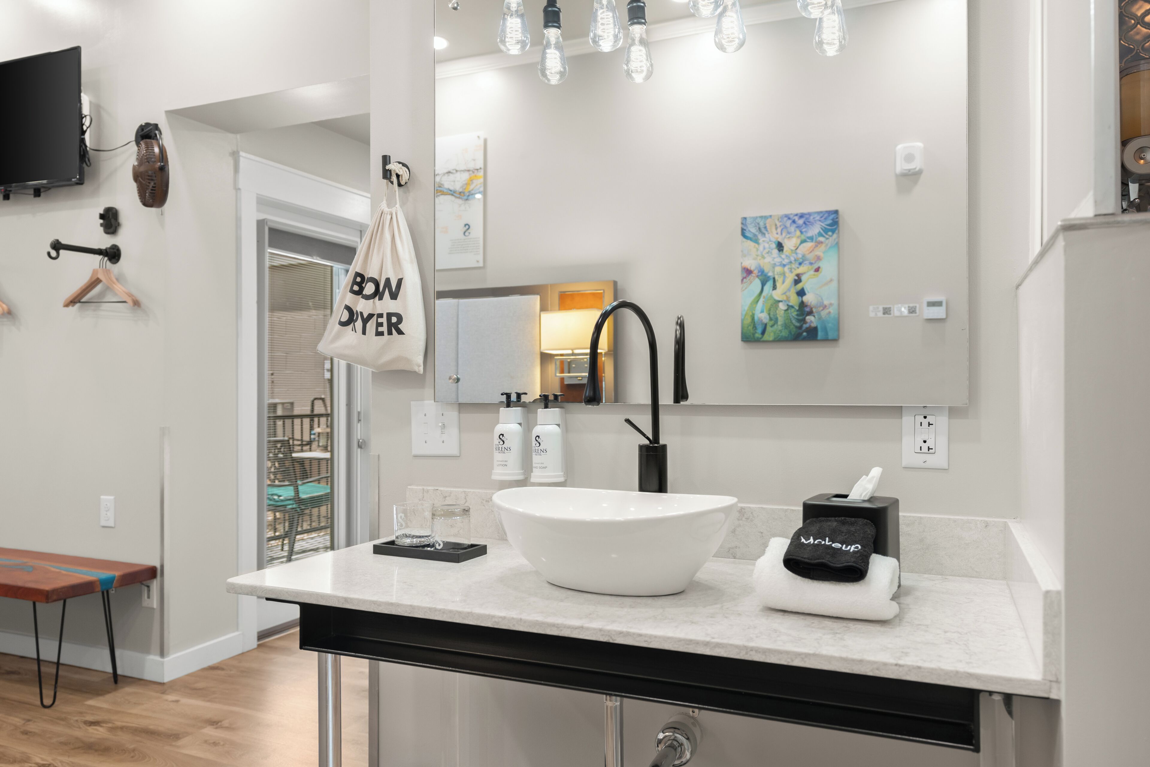 Ein modernes Badezimmer mit einem großen, weißen Waschbecken, einem schwarzen Wasserhahn und einem Spiegel darüber. An der Wand stehen zwei Lotionflaschen und ein Handtuch mit der Aufschrift 'makeup'.