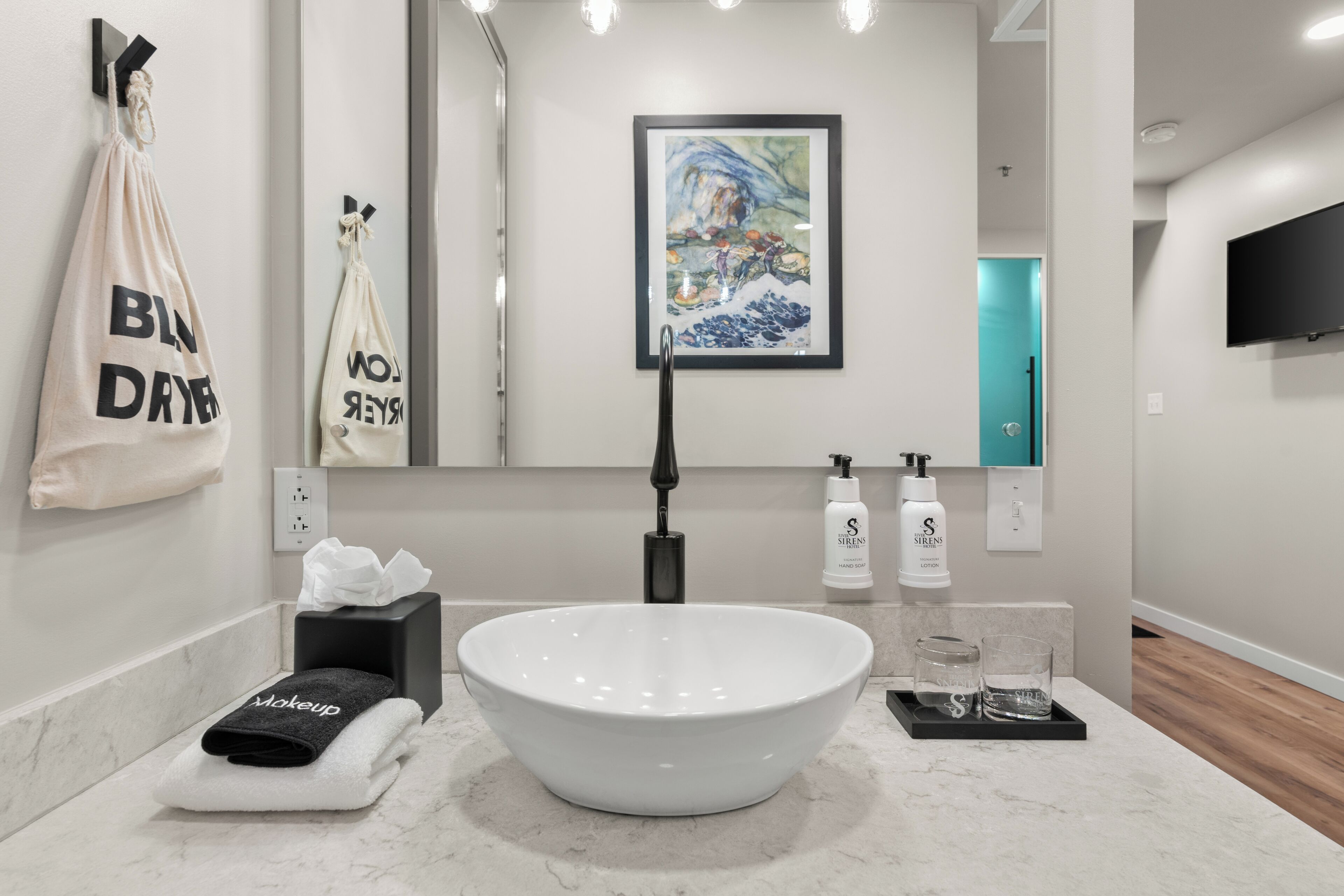 Une salle de bain moderne avec un grand lavabo blanc, un porte-serviettes muni d’un sac « BLN DRYER », une œuvre d’art encadrée et un téléviseur fixé au mur.