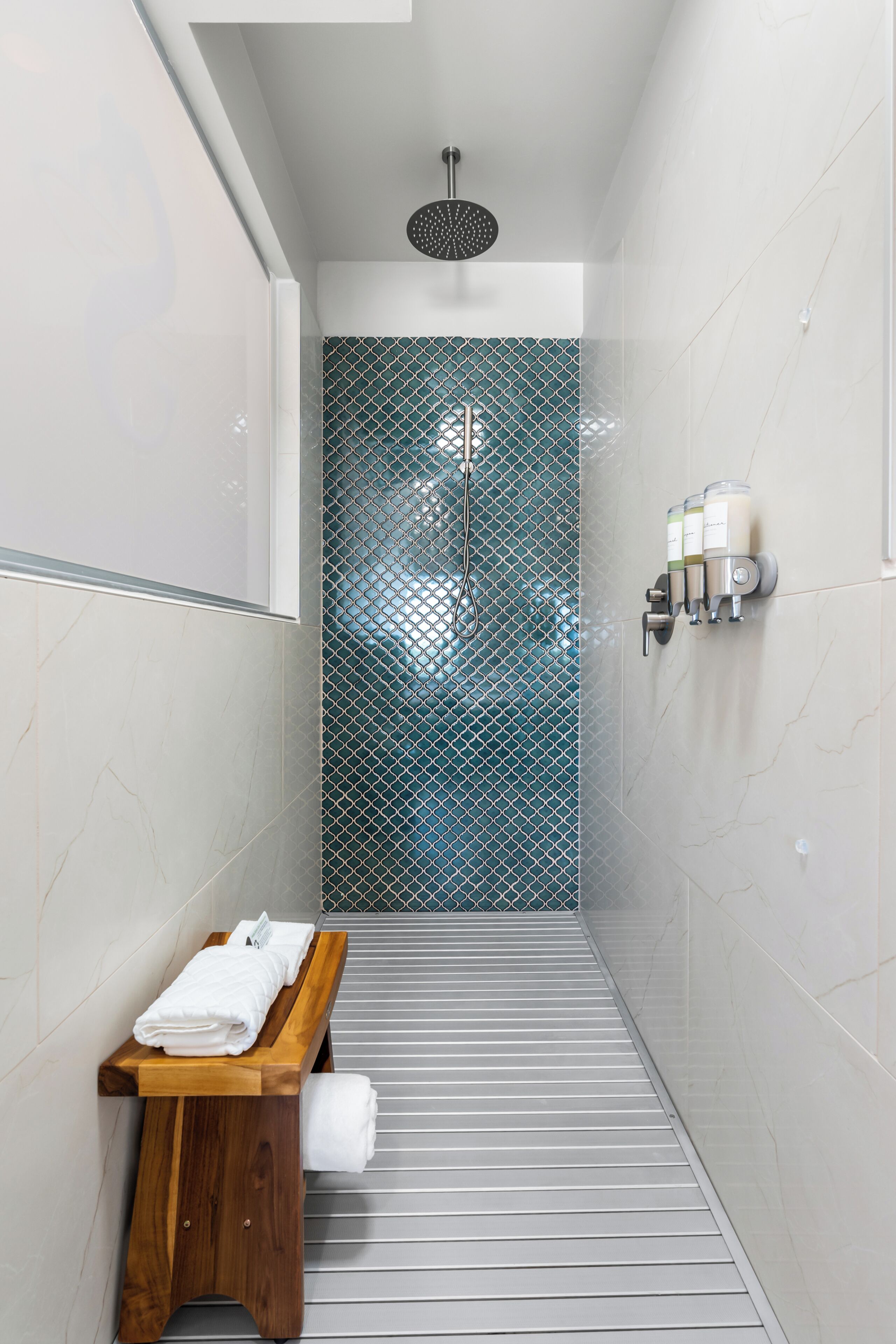 A modern shower with a glass door, a wooden bench with towels, and a wall-mounted soap dispenser.