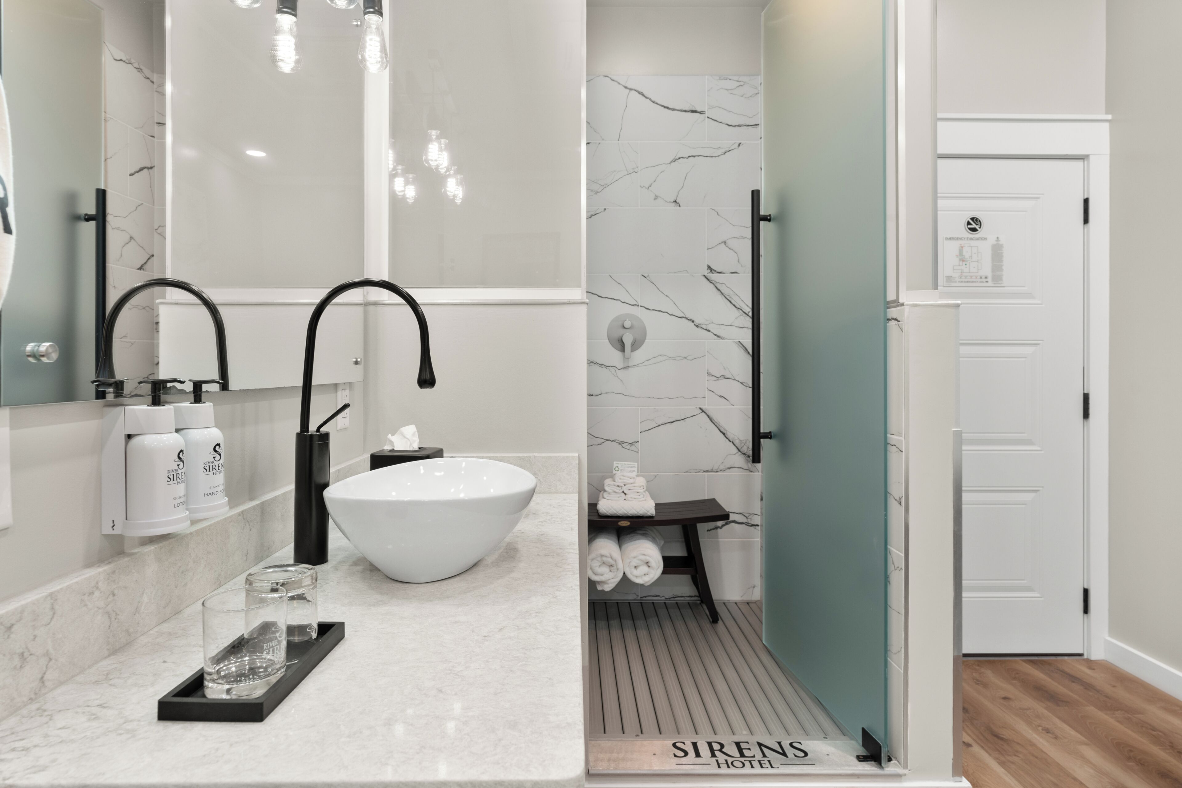 Un baño moderno con lavamanos blanco, grifo negro y azulejos de mármol. Hay artículos de aseo en el mostrador y un toallero con toallas dobladas.