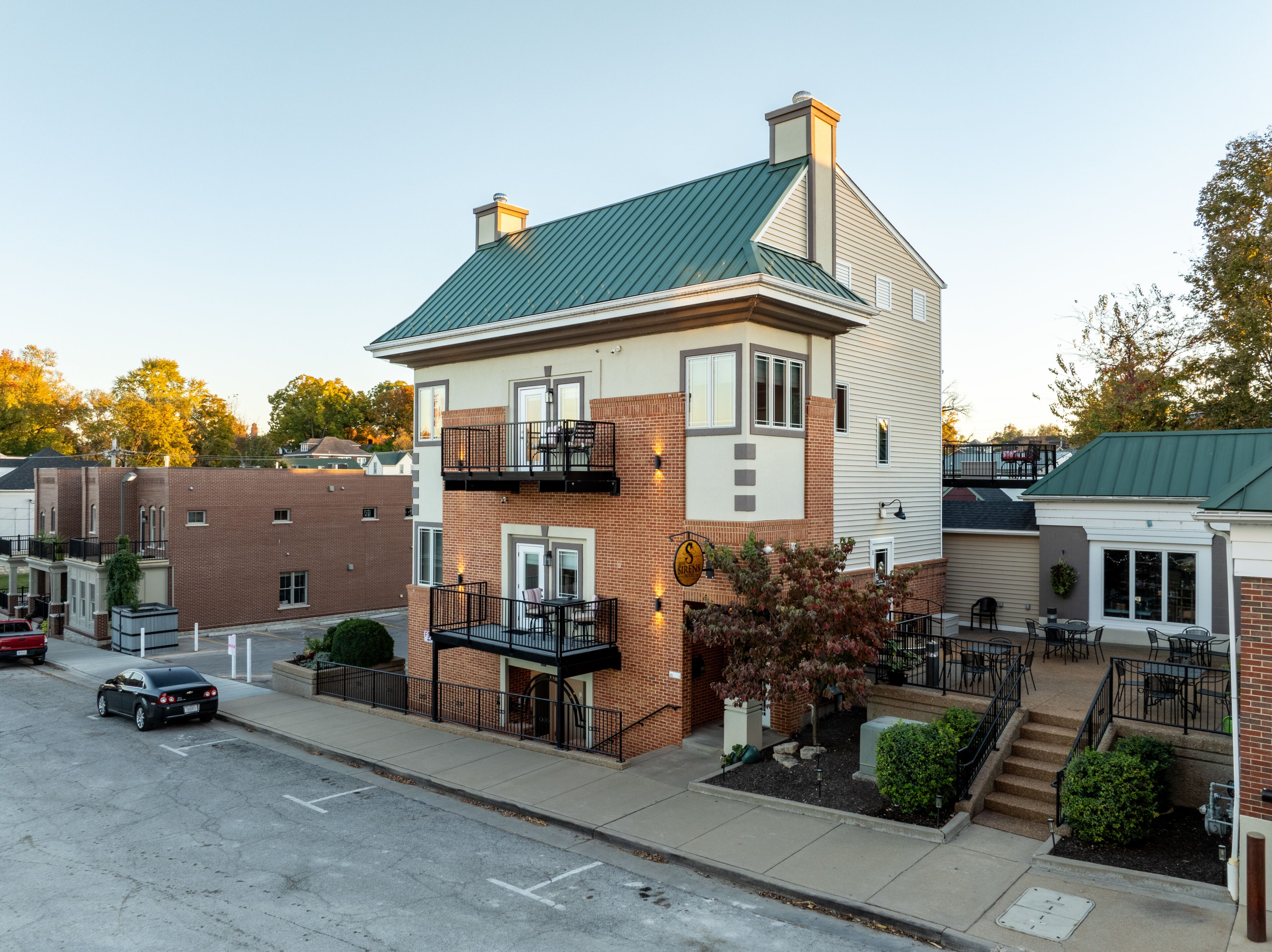 A two-story building with a green roof, a brick facade, and a small balcony with outdoor seating.