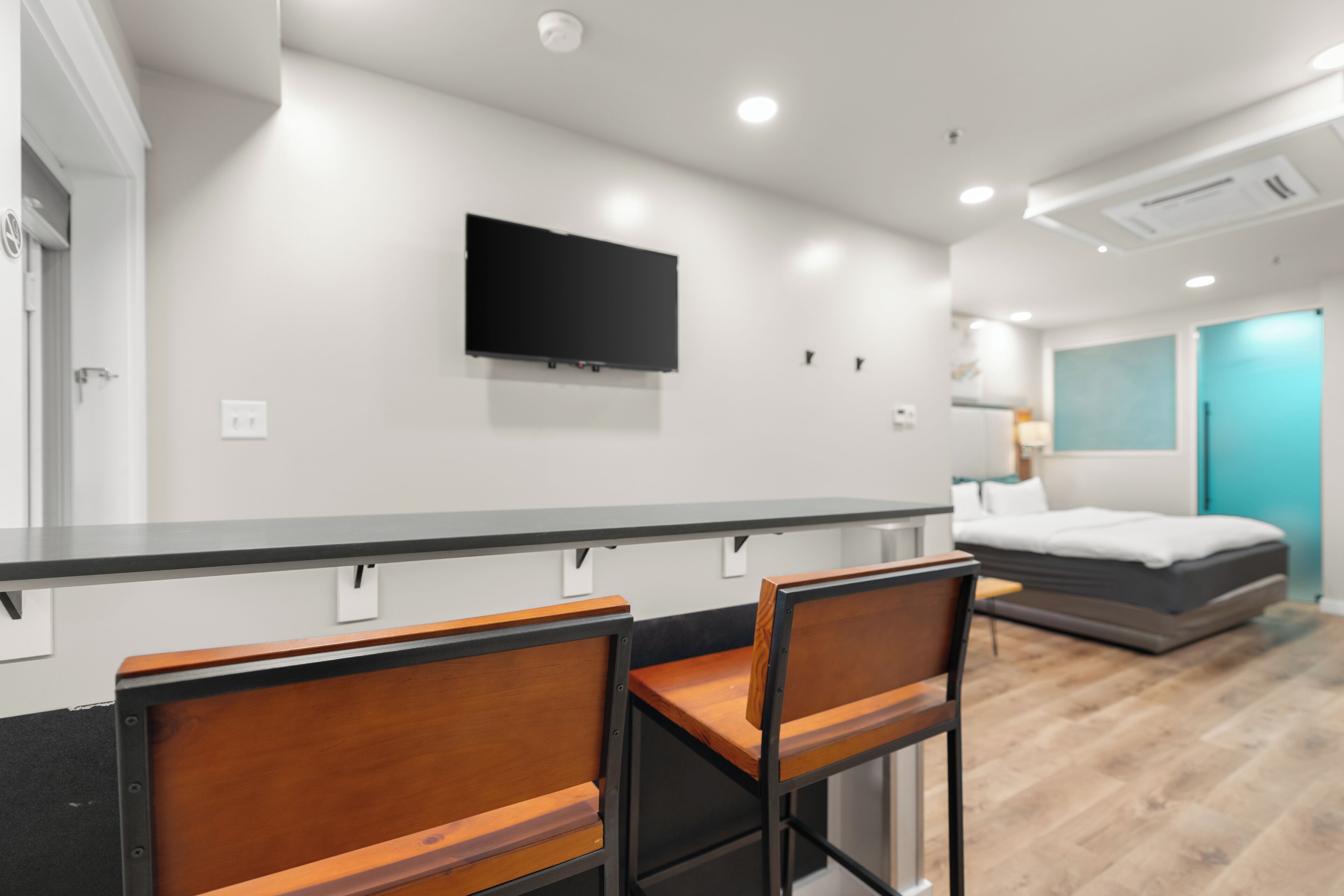Une chambre d’hôtel moderne équipée d’un téléviseur à écran plat fixé au mur, d’un bureau en bois, de deux chaises, d’un lit avec un matelas gris et d’une porte de couleur turquoise.