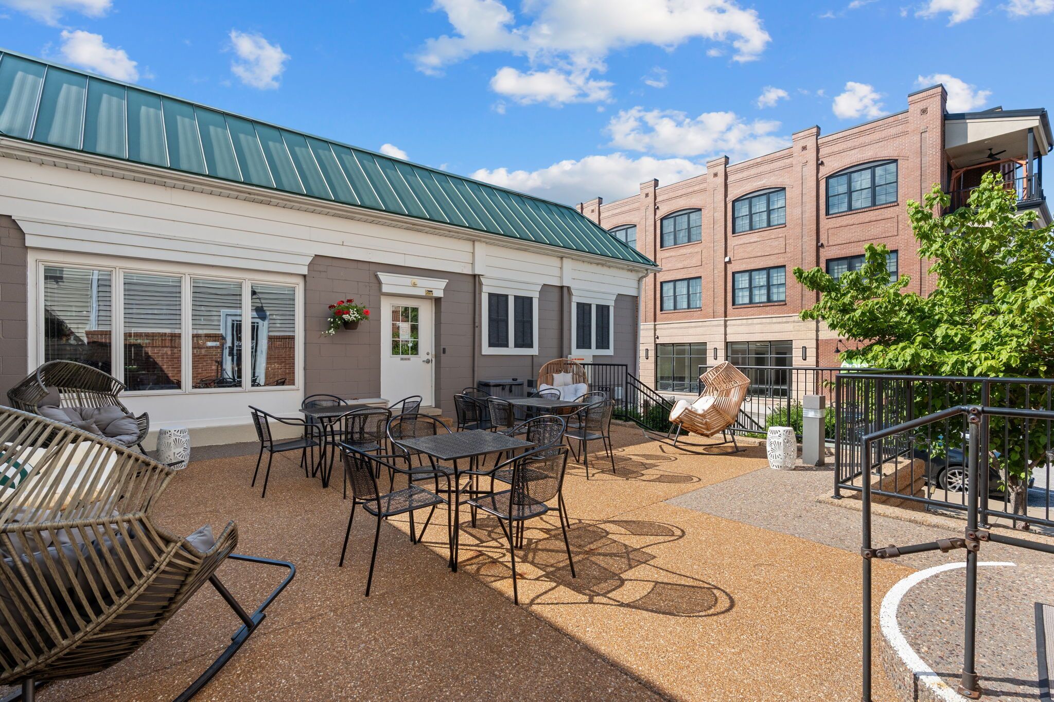 An outdoor patio with tables and chairs, a building with a green roof, and a brick structure in the background.