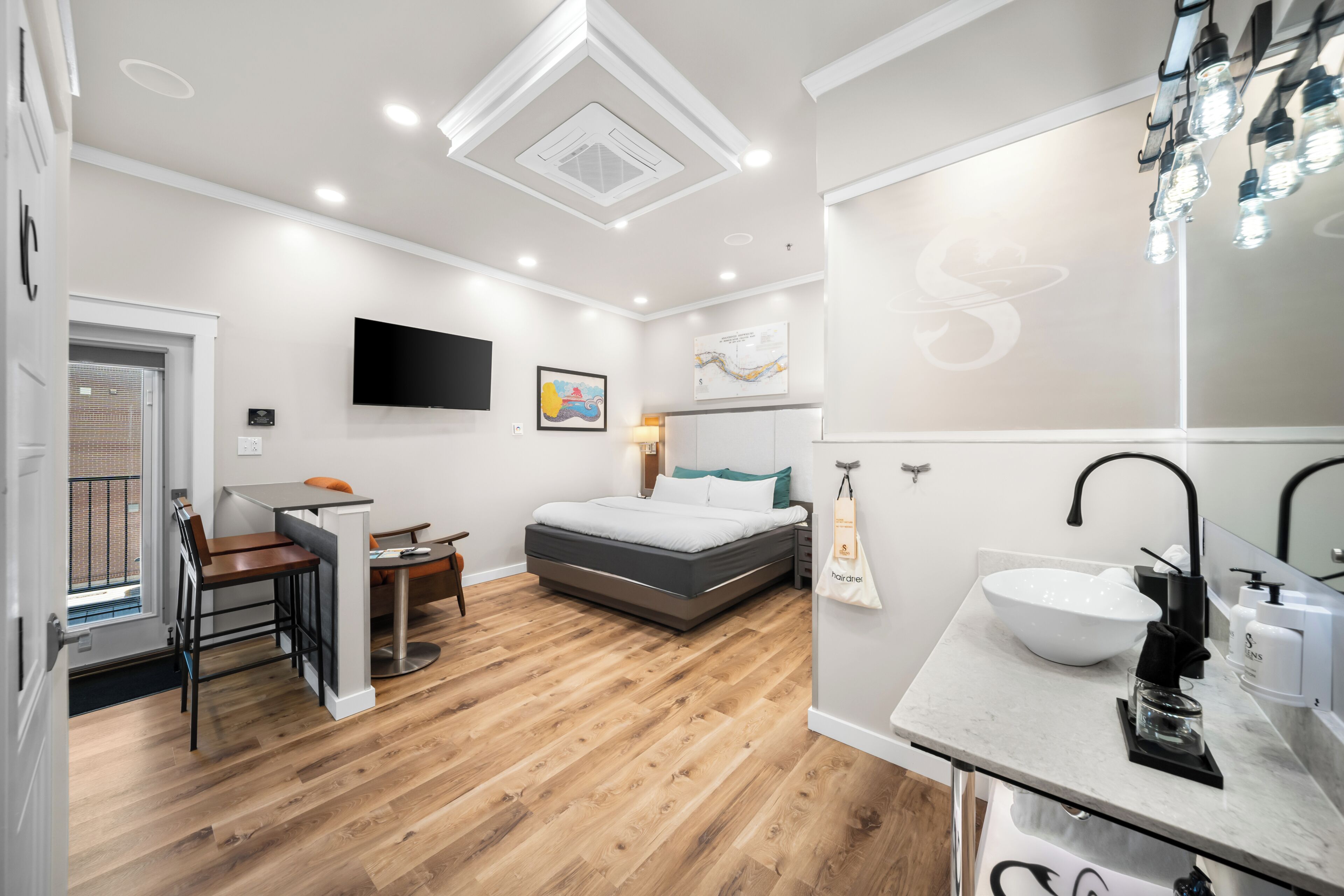 A modern hotel room with a bed, a desk, a chair, a TV, and a bathroom with a sink and mirror.