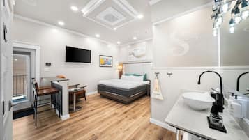 A modern hotel room with a bed, a desk, a chair, a TV, and a bathroom with a sink and mirror.