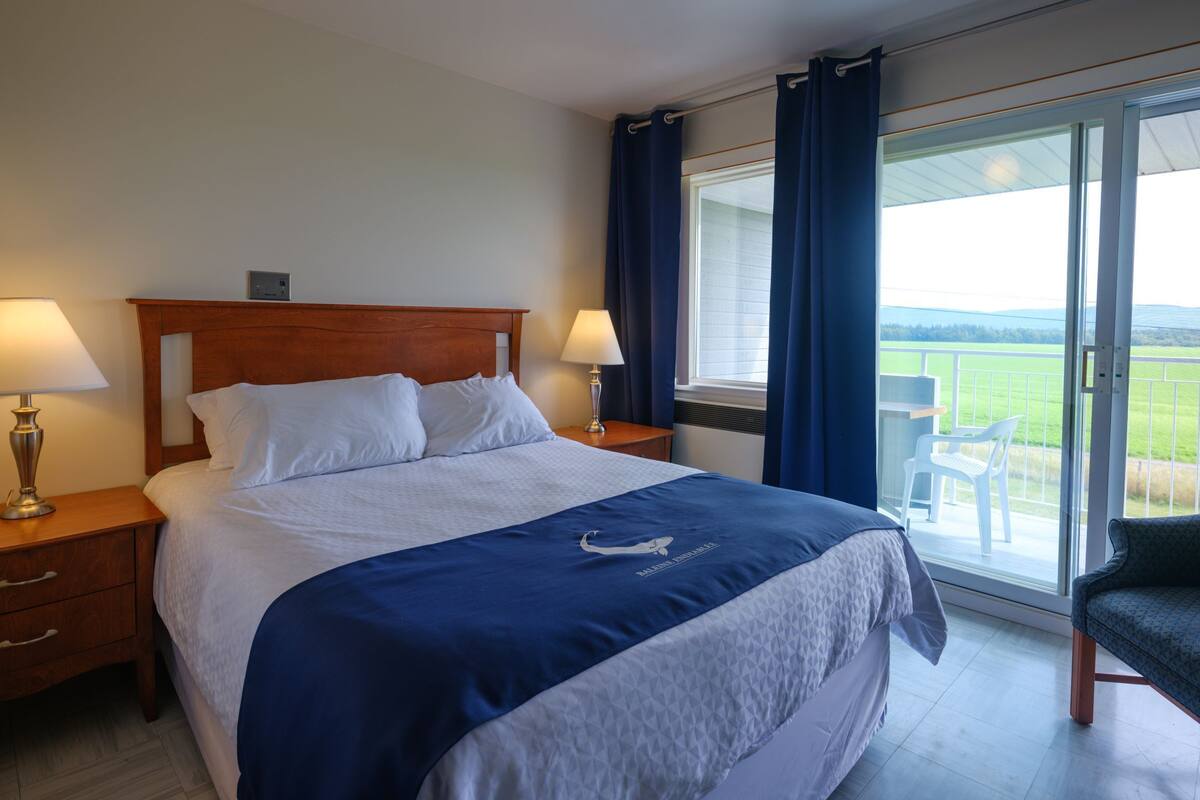 A hotel room with a large bed, two bedside tables with lamps, a window with curtains, and a view of the outdoors.