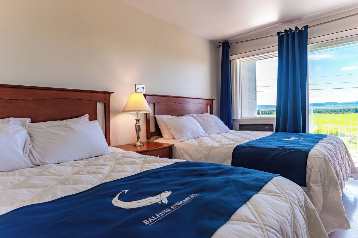 A hotel room with two beds, a nightstand with a lamp, a window with curtains, and a view of the outdoors.