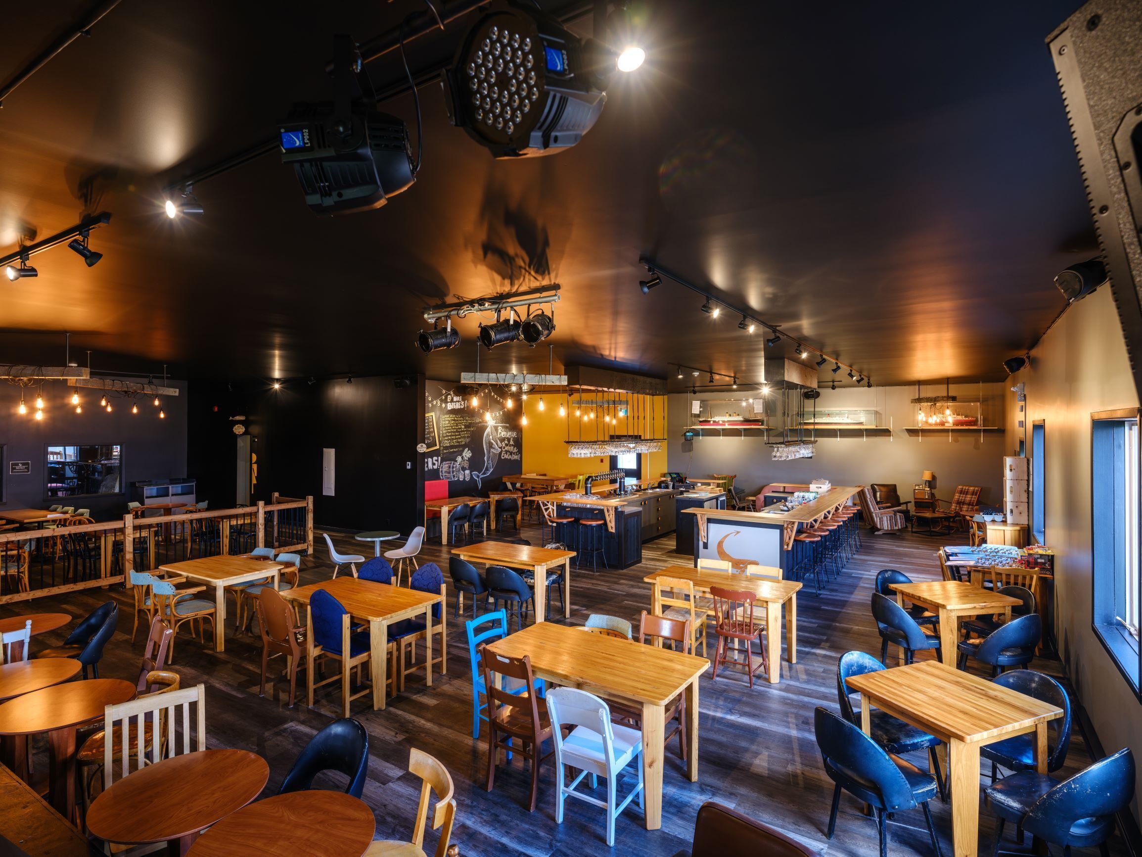 A restaurant with multiple wooden tables and chairs, a bar area, and a kitchen visible in the background.