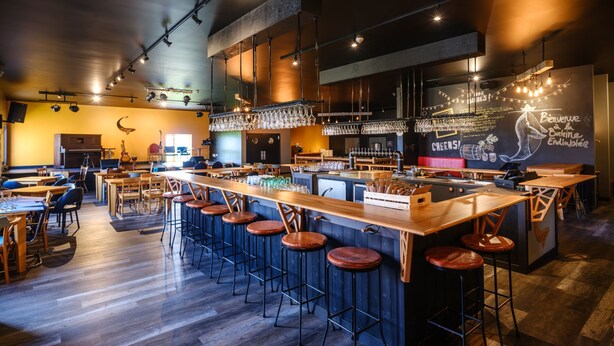 A modern bar with wooden stools, a long counter, and a chalkboard menu.