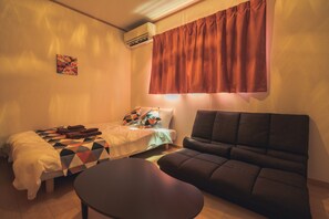 A small, cozy room with a bed, a sofa, and a small table. There is a window with curtains, an air conditioning unit, and a wall-mounted artwork. - Ferio101 (Tokyo)