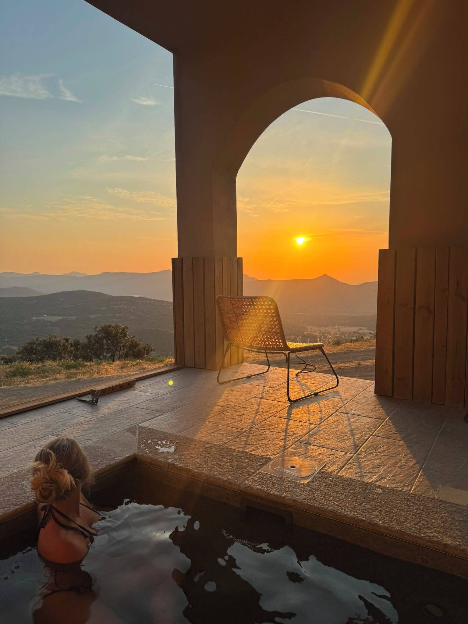 A sunset view from a covered outdoor seating area with a pool.