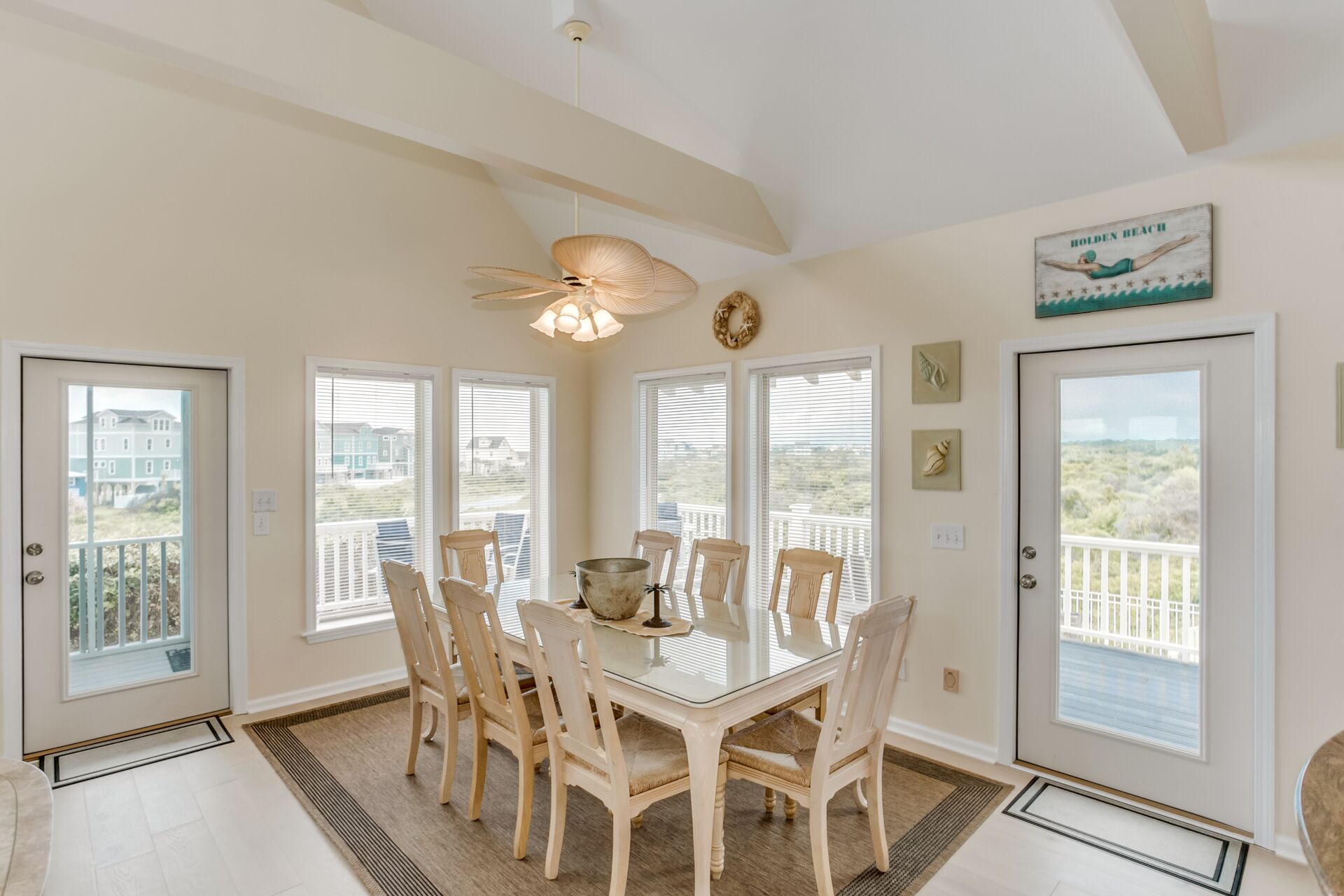 A dining room with a glass table surrounded by chairs, large windows with blinds, a ceiling fan, and a view of the beach.