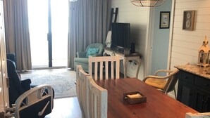 Uma área de refeições com mesa de madeira e cadeiras brancas, um ventilador de teto e uma televisão ao fundo.