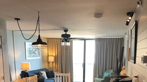 Uma sala de estar moderna com uma mesa de jantar de madeira, cadeiras brancas e um sofá. Há também uma área de cozinha com bancada e pia.