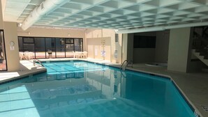 Piscina com água azul cristalina, rodeada por um deck revestido e uma área coberta com cadeiras.