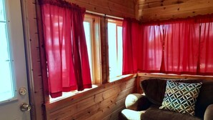 A wooden cabin interior with a sofa and a patterned pillow, red curtains, and a door with a handle.