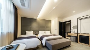 A modern hotel room with two beds, a small desk, a chair, and a compact kitchenette.