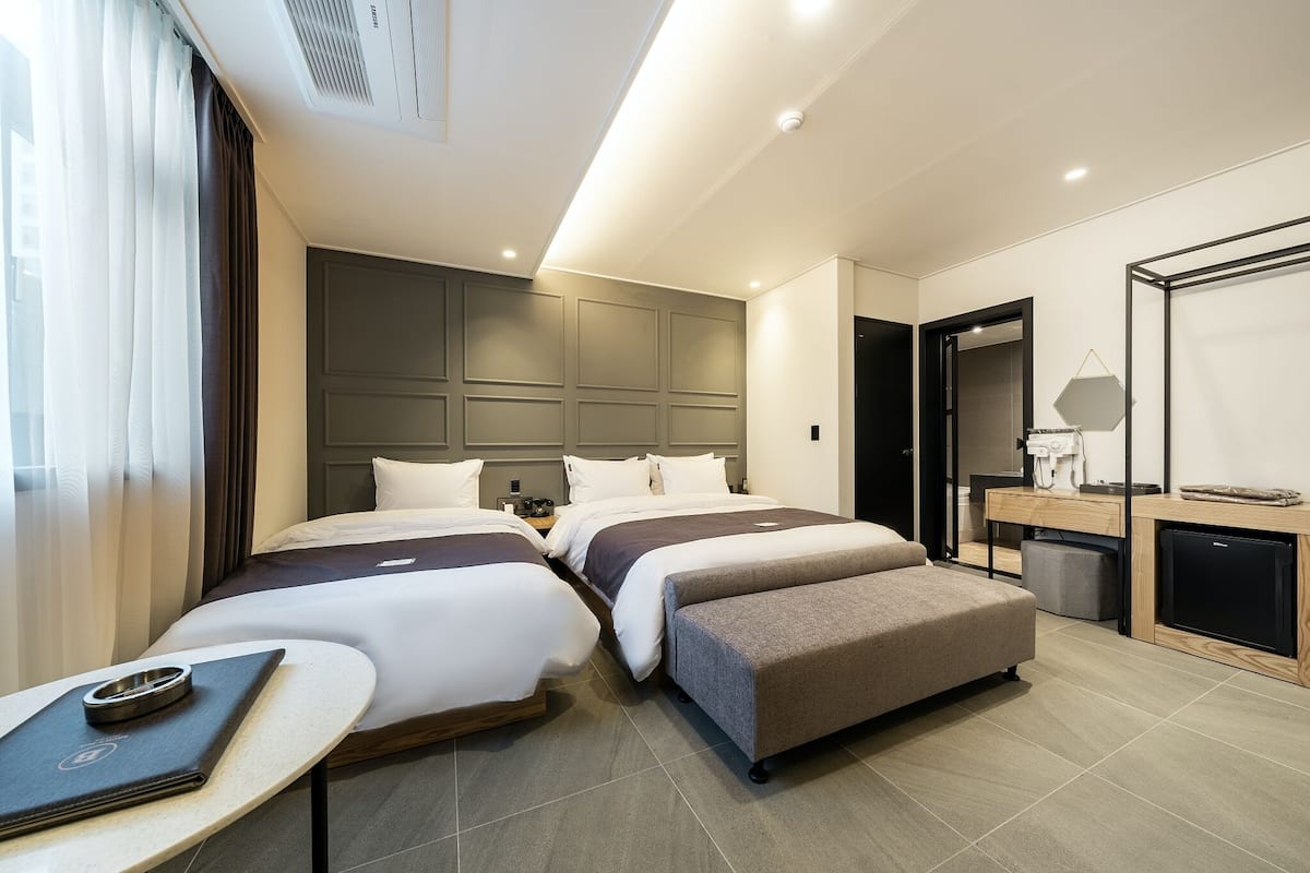 A modern hotel room with two beds, a small desk, a chair, and a compact kitchenette.