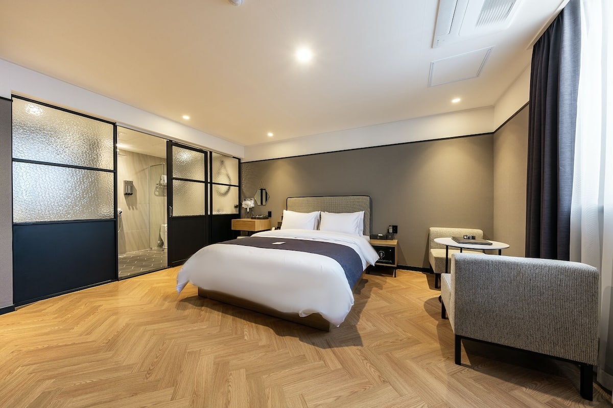 A modern hotel room with a large bed, a seating area, a small table, and a glass-enclosed shower.