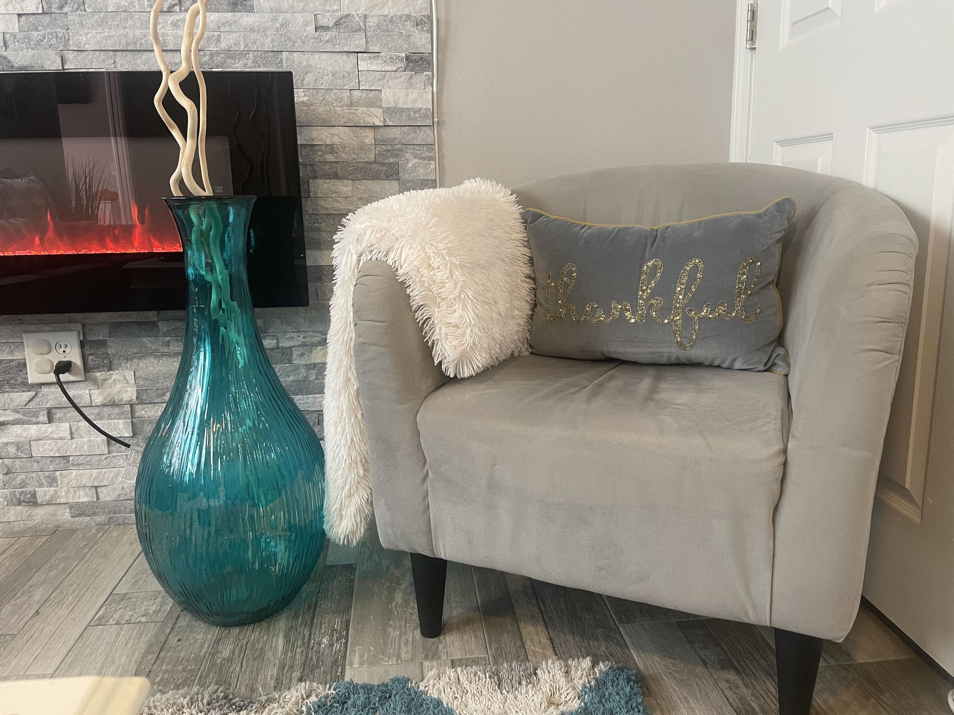 Ein Wohnzimmer mit einer türkisfarbenen Vase, einem grauen Sofa mit weicher Decke und einem Kissen mit der Aufschrift 'dankbar'.