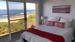 A bedroom with a large sliding glass door leading to a beach view, a bed with white bedding and a red throw, and a painting on the wall.