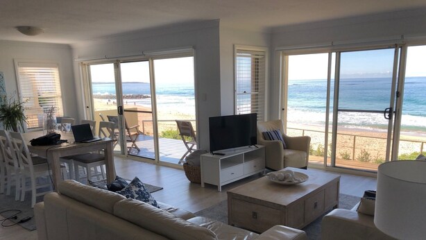 A modern living room with a sofa, coffee table, TV, and a balcony with a view of the beach and ocean.